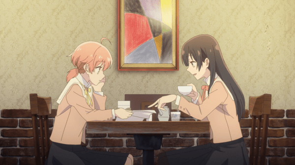 Two teen girls sit at a table in a coffee shop, looking at a set of papers in one girl's hand.